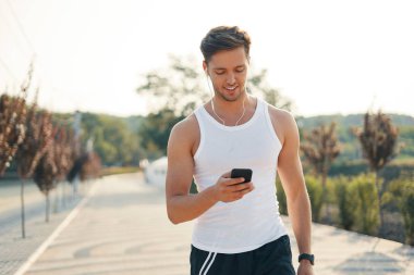 Çekici, gülümseyen, sportif genç adam onun müzik başlamak üzere kadeh çalma listesi üzerinde smartphone için ekran açık havada bakarken.