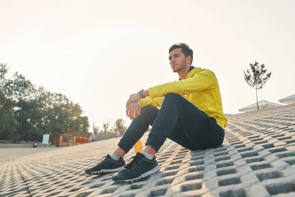 Sports. Athletic Man With Fit Body In Yellow Sportswear Resting on Gray Grunge After Running, Jogging Outdoors. Handsome Healthy Male Runner Taking Break After Fitness Workout And Exercising.