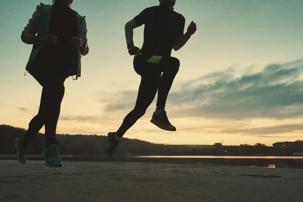 年轻的夫妇跑步者的腿在日出时跑 背景是湖和天空 运动的人在户外训练 向下角度拍摄 — 图库照片