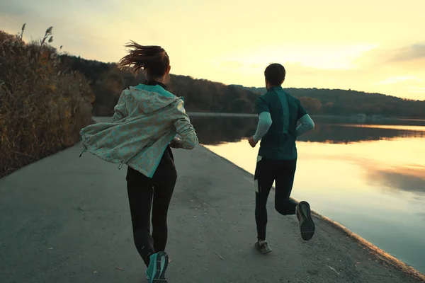 Motivated couple of runners out for a run on the lake at the sunrise. Young man and woman in sport clothing, running together. Sportive people training outdoors