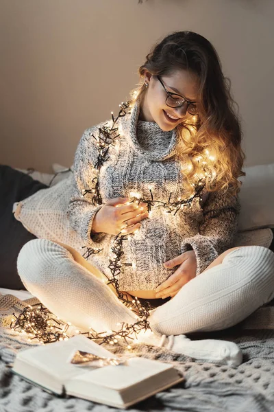 Rahat, yumuşak kazak, uzun beyaz çorap ve elbiselerini üzerinde sarı ışıklı gözlük gülümseyen hamile genç kadın eve yatağa süre oturma rahatlatıcı bacak, bacak. 