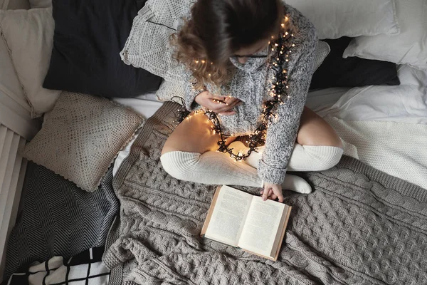 Hamile kadının ellerini karnına dokunmak ev rahatlatıcı, yatağa oturmuş bir kitap bacak bacak okuma, elbiselerini üzerinde sarı ışık ile rahat, yumuşak kazak, uzun beyaz çorap giyen süre