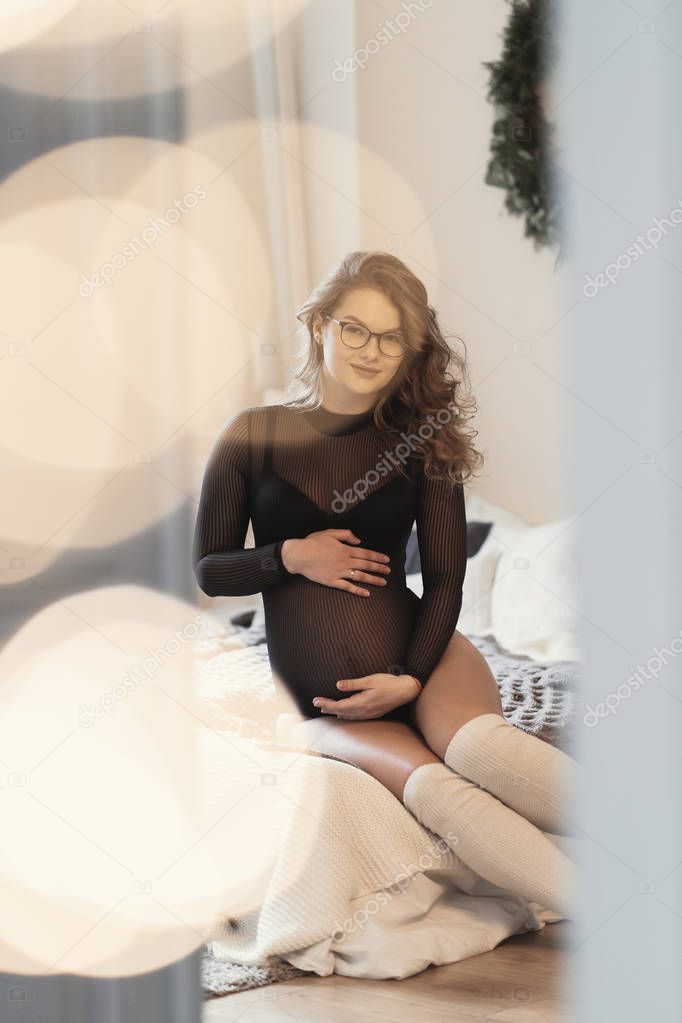Beautiful pregnant woman, wearing transparent black body lingerie and white long socks enjoying her feminine body while touching her belly sitting on bed at home. Shot through door with bokeh lights 