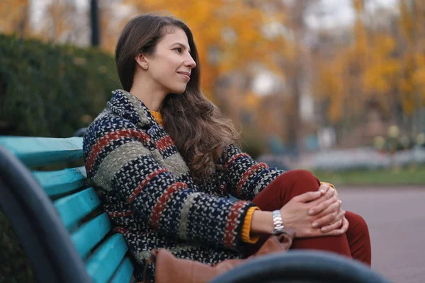 Pretty young woman in good mood posing in the autumn day, while sitting on a bench, enjoying the good weather.