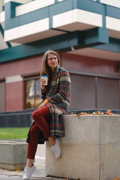 屋外コーヒーを飲みながらコートでスタイリッシュな若い女性 現代都市の服装と休憩をしながら街の通りに座っているメガネのカジュアルな女の子 — ストック写真