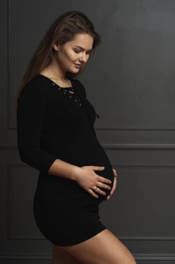 Siyah elbise giymiş, yavaşça karnı elleriyle dokunuyor, doğmamış bebeğini gri duvar arka plan üzerinde poz için aşk gönderme muhteşem genç hamile kadın portre,.