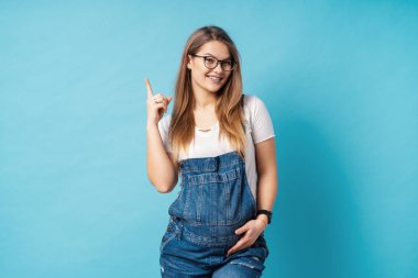 Harika bir fikir kadar işaret parmağı ile mavi arka plan üzerinde gösteren gözlük ve denim romper genç ve güzel hamile kadın.