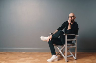 Bacak bacak, kendinden emin bir poz Studio bir gri duvar arka plan, gümüş bir iskemle üzerinde oturan bir modern, şık adam. O sakalı, Gözlük, siyah rahat kıyafetler ve beyaz spor ayakkabı giyiyor.