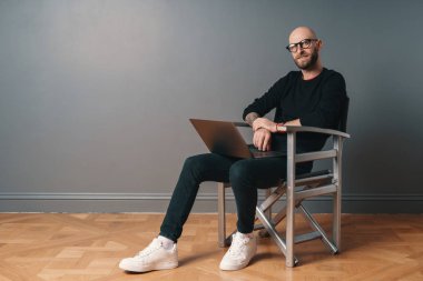 Sakal ve studio iç rahatlatıcı, bir gümüş sandalye, gri arka plan üzerinde oturan ve dizüstü bilgisayarda internet sörf gözlük modern beyaz adam. Gadget'ı kullanarak çevrimiçi iletişim zevk adam.