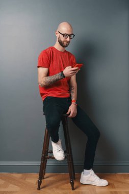 Yakışıklı tüysüz beyaz adamla sakal, Gözlük, kırmızı T-shirt telefonda konuşurken, smartphone ekranında, gri studio arka plan üzerinde bakarken dövmeli kollarında tutarken manifatura.