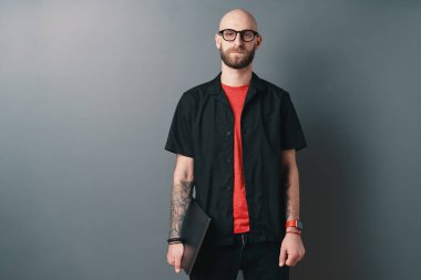 Genç bir adam gözlüklü gri arka plan üzerinde Studio dizüstü kol altında tutan sakallı. Şık tasarımcı, iyi bir tasarım deneyimi onun müşterilerine sunuyor.