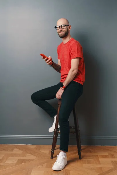 Handsome hairless Caucasian man with beard, glasses, red T-shirt texting while holding smartphone in his tattooed arms, smiling, he is posing while sitting on a tall chair, over gray studio background
