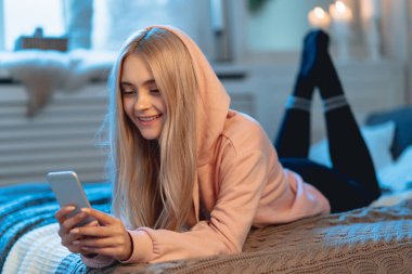 Sarışın kız pembe parka ve arkadaşları ile sohbet ederken evde, yatakta yatıyordu bir akıllı telefon kullanarak siyah tozluk gülümseyen