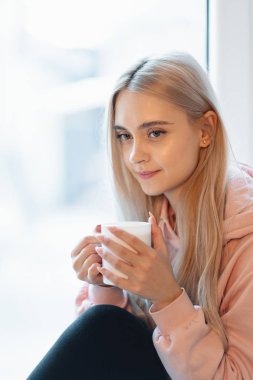 Bir fincan kahve ya da süt bulanık pencere cam tarafından otururken tadını çok gülümseyen bir kız. Rahat bir pembe, soluk parka giyiyor. Dudaklar yakınındaki kadın elinde beyaz kupa kapatın.