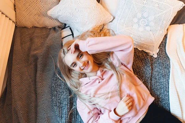 Beautiful smiling girl, with long blond hair, enjoying happiness, she cant belive its true. Portrait gorgeous young woman lying on the bad with pillows in comfy pink pale parka looking at camera.