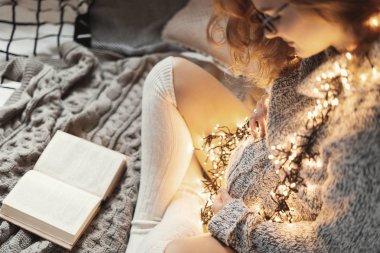 Evde oturan yatağa rahatlatıcı giyiyor rahat, bacak, bacak iken karın dokunmadan hamile kadının elleri yumuşak kazak, uzun beyaz çorap, elbiselerini üzerinde sarı ışık ile. Kırpılmış görüntü