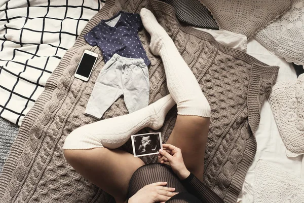 Expecting pregnant woman touching her belly while looking at ultrasound scans, sitting on cozy bed with blankets, pillows, kid clothes, smartphone, wearing white long socks. Cropped image top view.