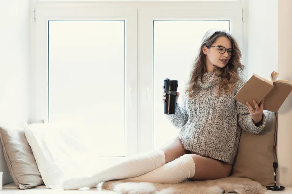 Relaxed beautiful pregnant woman dreaming while sitting on the window sill with bokeh light around reading a book and drinking tea from a thermos. She wear a warm, soft sweater and long white socks.