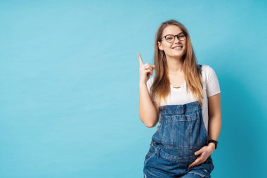Mavi arka plan üzerinde büyük bir fikir kadar indeks parmak ile işaret güzel genç hamile kadın