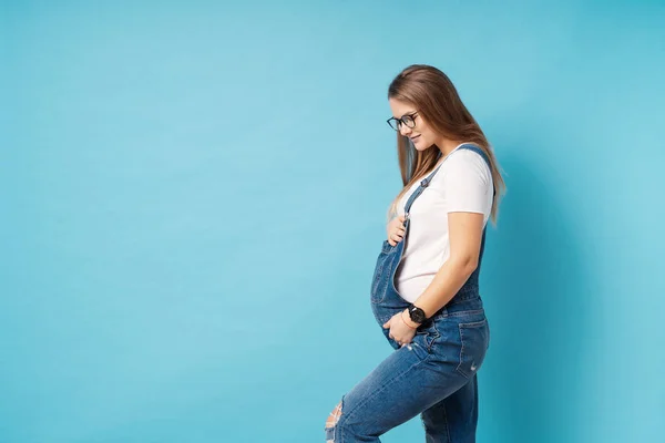 Usmívající se těhotná žena, která nosí brýle a hladil si břicho na modrém pozadí — Stock fotografie