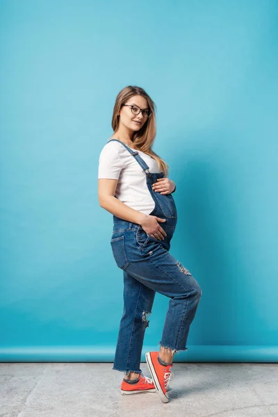 Sourire femme enceinte portant des lunettes caressant son ventre sur fond bleu — Photo