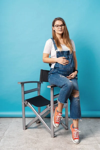 Gravid kvinna smeker hennes mage medan du sitter på en hög stol i studion över blå bakgrund. — Stockfoto