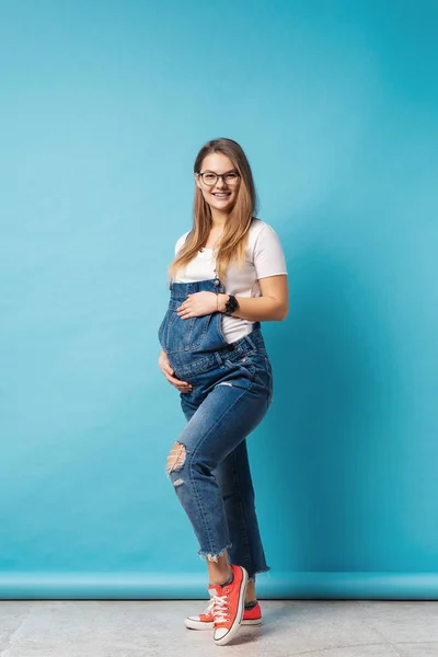 Sorridente donna incinta indossa occhiali accarezzando la pancia su sfondo blu — Foto Stock