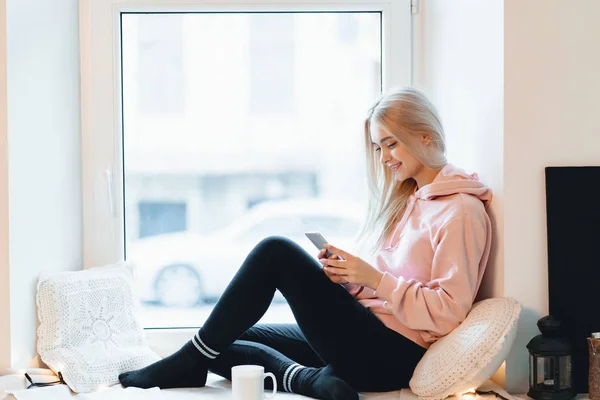 Όμορφη νεαρή γυναίκα που απολαμβάνει καφέ ενώ χρησιμοποιεί ένα smartphone από τα παράθυρα σε εσωτερικούς χώρους — Φωτογραφία Αρχείου