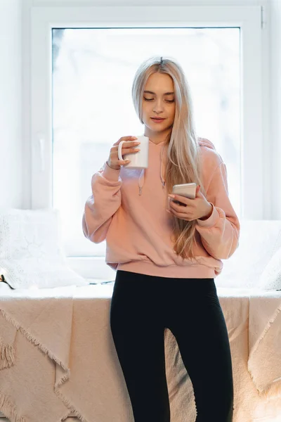Mooie jonge vrouw genieten van koffie terwijl het gebruik van een smartphone door de ramen binnenshuis — Stockfoto