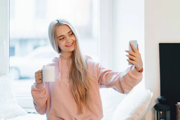 Gondtalan lány így selfie egy csésze kávé mellett állva az ablak közelében otthon — Stock Fotó
