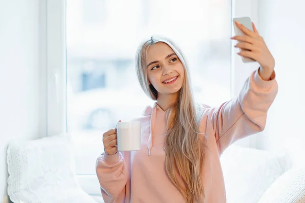 Menina despreocupada fazendo selfie com uma xícara de café enquanto está perto da janela em casa — Fotografia de Stock