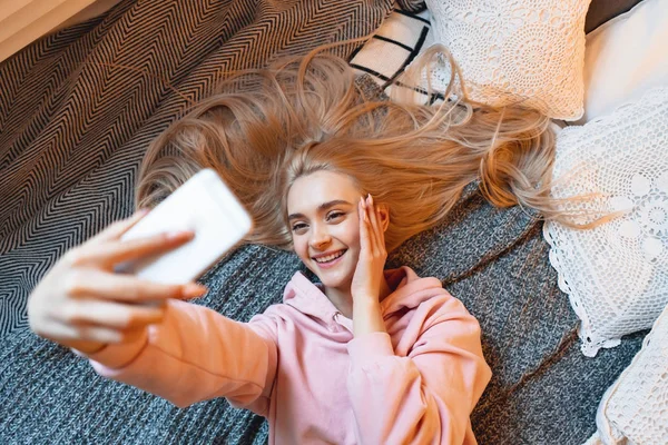 Güzel genç kız evde yatakta yatarken akıllı telefon ile selfie yapıyor, eğleniyor — Stok fotoğraf