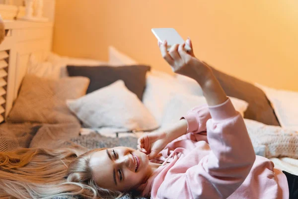 Красивая молодая девушка развлекается, делает селфи со смартфоном, лежа на кровати дома — стоковое фото