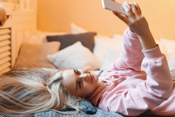 Ładna młoda dziewczyna zabawy, co selfie z inteligentnym telefonem leżąc na łóżku w domu — Zdjęcie stockowe