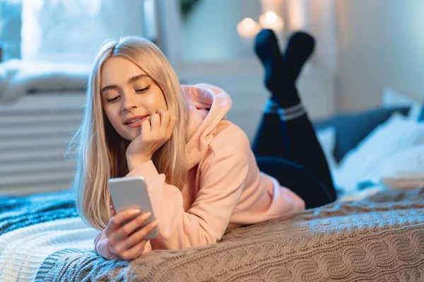 Szép szőke lány rózsaszín segítségével okos telefon feküdt az ágyon otthon, míg beszélgetni a barátaival — Stock Fotó