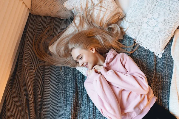 Улыбающаяся девушка, с длинными светлыми волосами, наслаждающаяся счастьем, лежа на кровати дома — стоковое фото