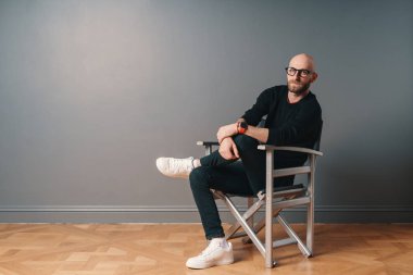 Kendine güvenen bir poz oturan modern bir adam, bir sandalyede bacak üzerinde bacak, stüdyoda gri duvar arka plan üzerinde