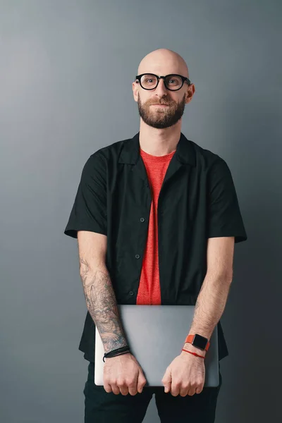Ответственный бородатый мужчина в очках, держащий ноутбук под рукой в студии на сером фоне — стоковое фото