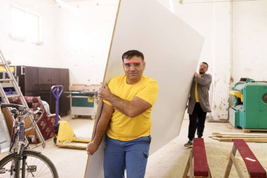 İki adam atölyelerinde mobilya malzemeleri taşıyor.