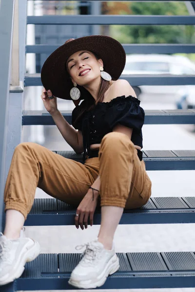 Mulher morena elegante com cabelo longo, vestindo chapéu de vime grande elegante, posando nas escadas — Fotografia de Stock