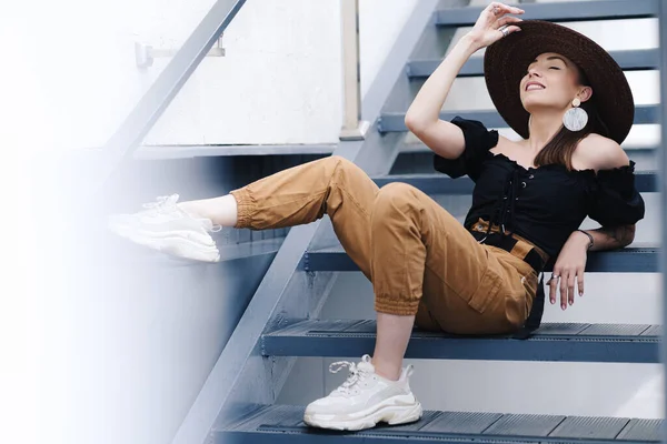 Mujer morena de moda con el pelo largo, con elegante sombrero de mimbre grande, posando en las escaleras —  Fotos de Stock