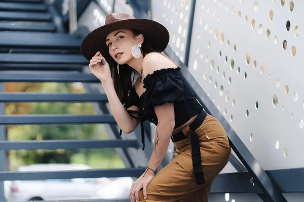 Divatos barna nő hosszú hajjal, elegáns nagy fonott kalapot visel, pózol a lépcsőn — Stock Fotó