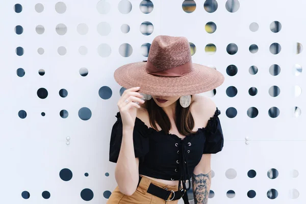 Modieuze brunette vrouw met lang haar, het dragen van stijlvolle grote rieten hoed, poseren tegen een witte muur met gaten — Stockfoto