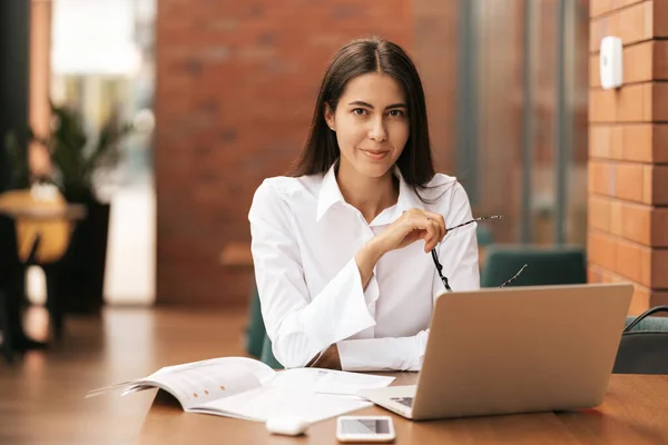 Hermosa mujer en gafas hablando con el ordenador portátil con sonrisa mientras está sentado y trabajando — Foto de Stock