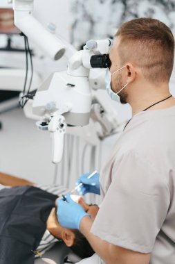 Erkek dişçi maske takıyor. Mikroskoba bakıyor.