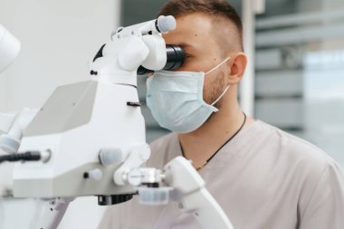 Erkek dişçi maske takıyor. Mikroskoba bakıyor.