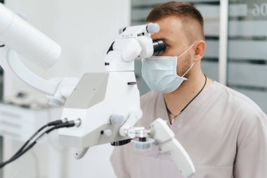 Erkek dişçi maske takıyor. Mikroskoba bakıyor.