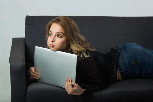 beautiful young girl lying on a black sofa with a laptop on a white background