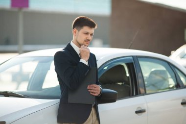yakışıklı genç düşünen adam bir takım elbise ayakta yakınındaki bir klasörle açık havada beyaz bir araba