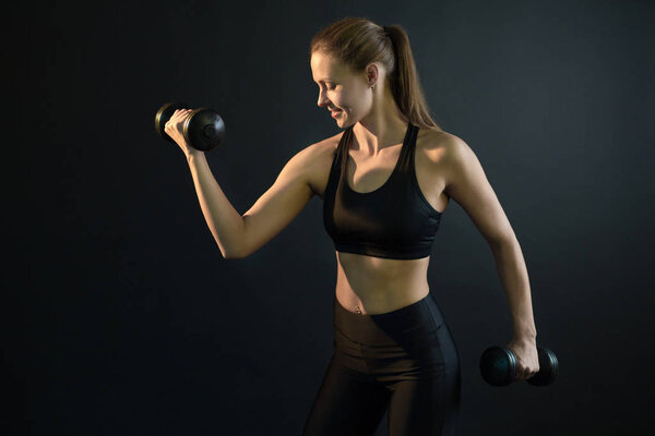 beautiful young girl training with dumbbell shaking her biceps on a black background